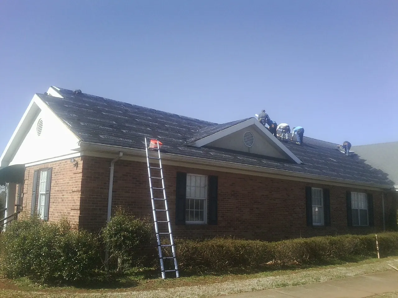Crew tearing shingles off a brick ranch roof with a ladder set — Emergency Roof Repair work by our crew in Chesapeake