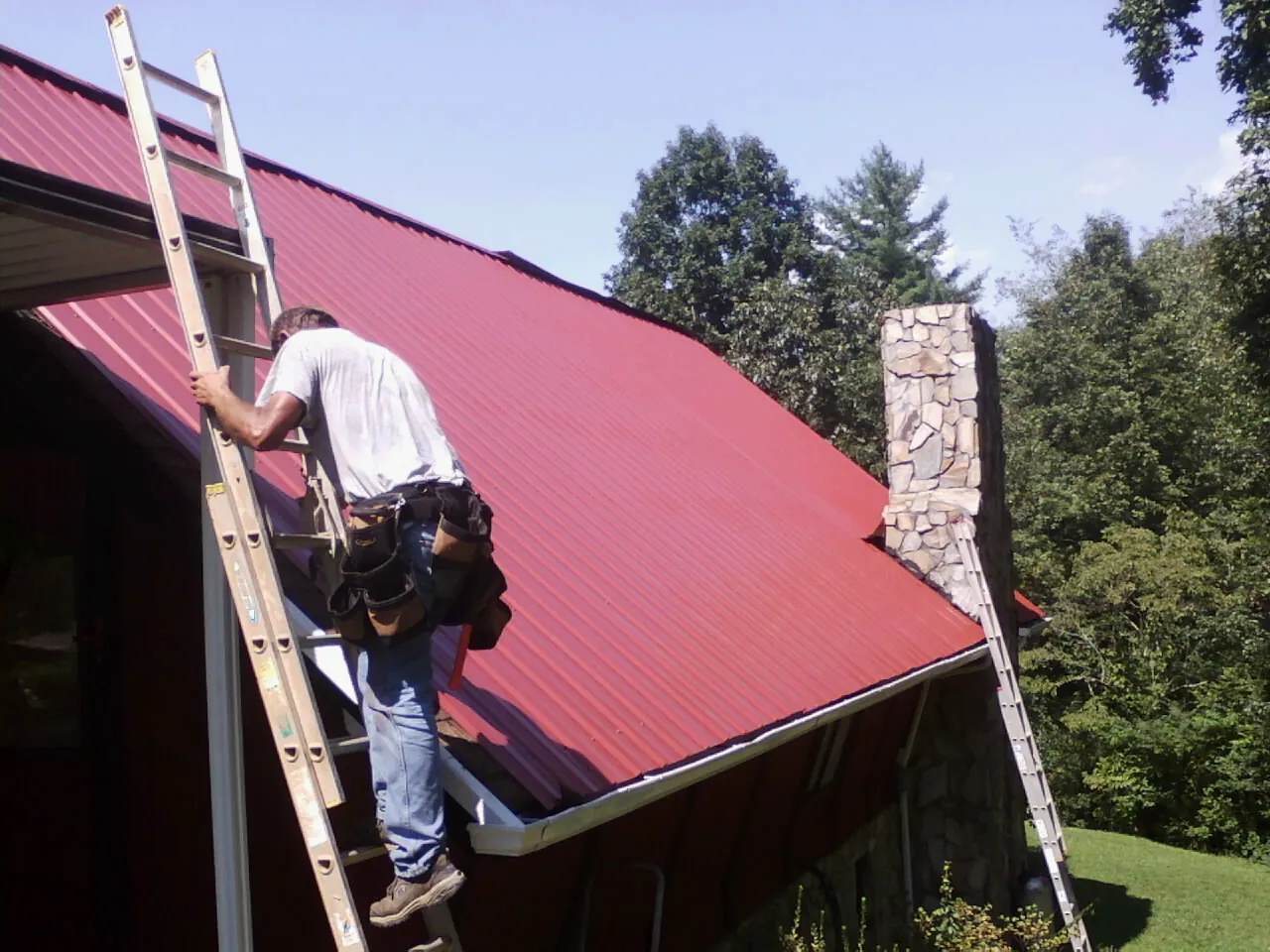 Roofer on a ladder beside a red metal roof and stone chimney — Soffit & Fascia Repair by Roofing Brothers in Chesapeake